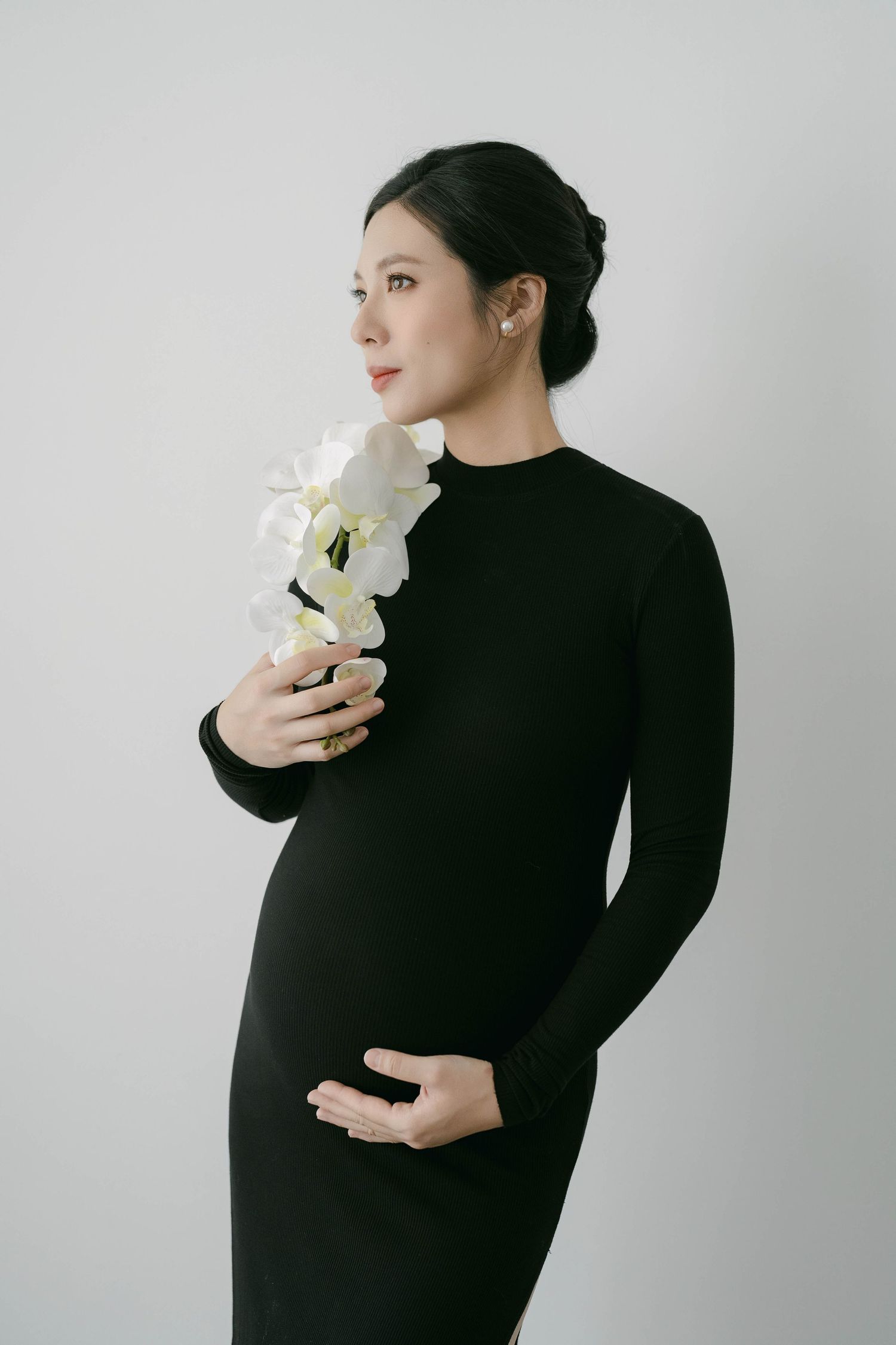Maternity photoshoot in black dress with white flowers against neutral background.