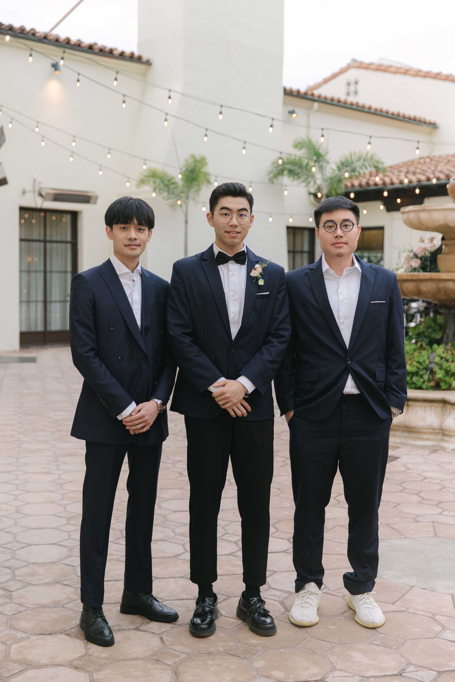 Three men in formal suits standing together at an outdoor courtyard with string lights.