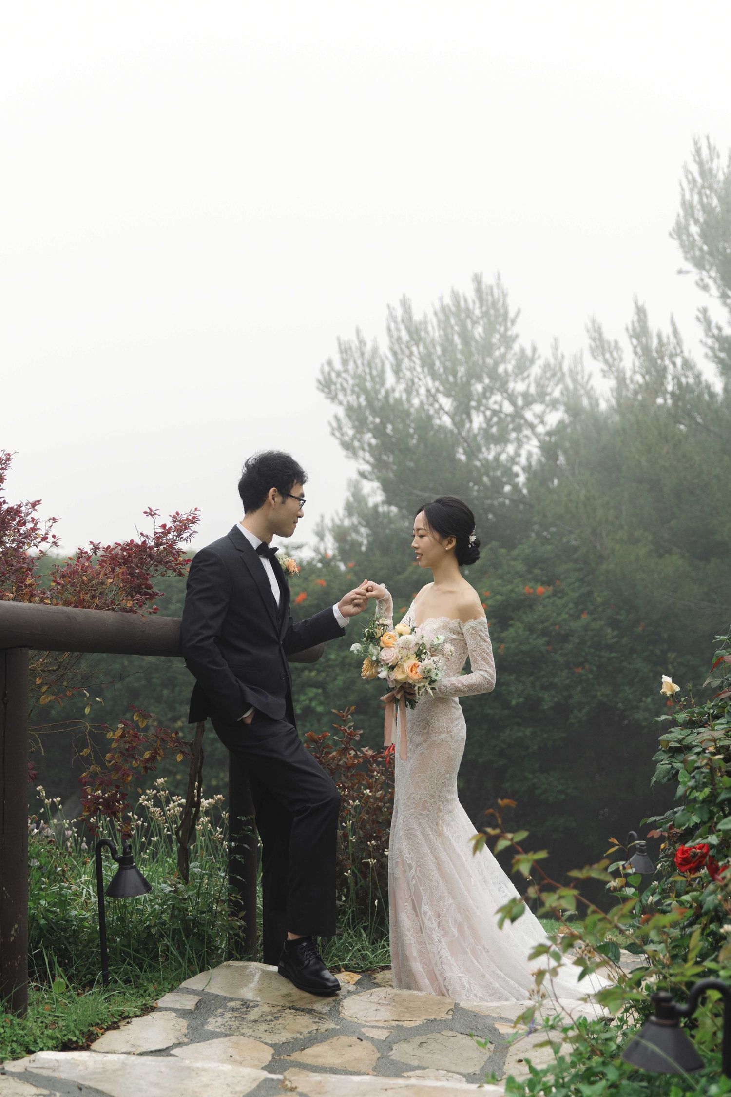 Newlyweds exchange loving glances while holding bouquets in a misty garden setting.