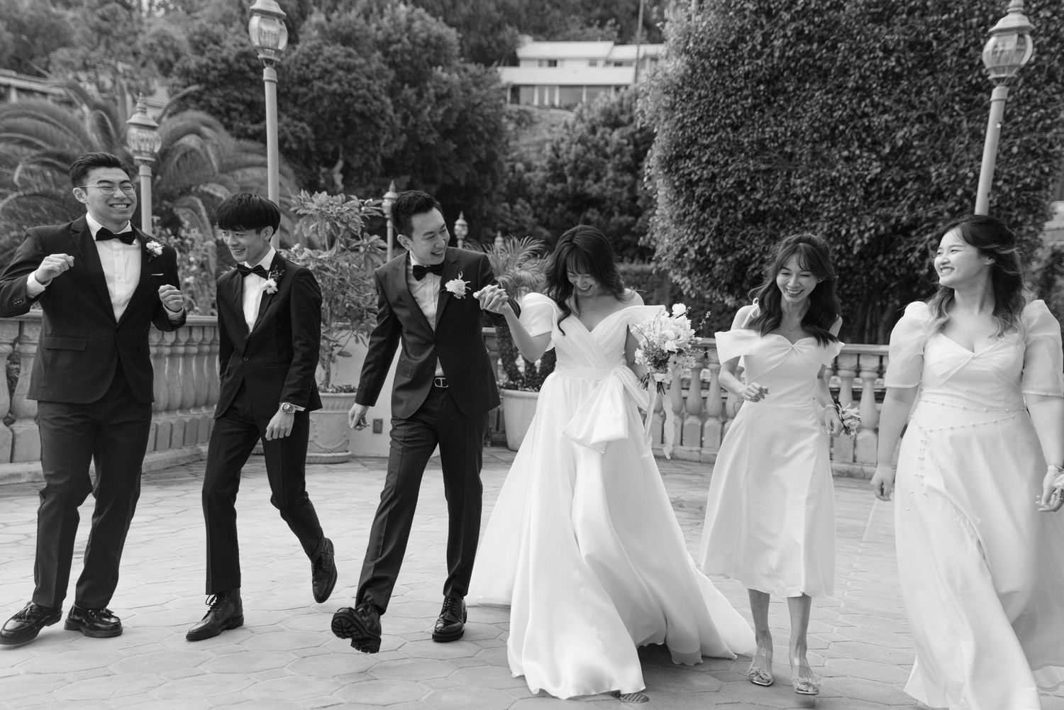 Wedding party members in formal attire walk in a line on a stone pathway.