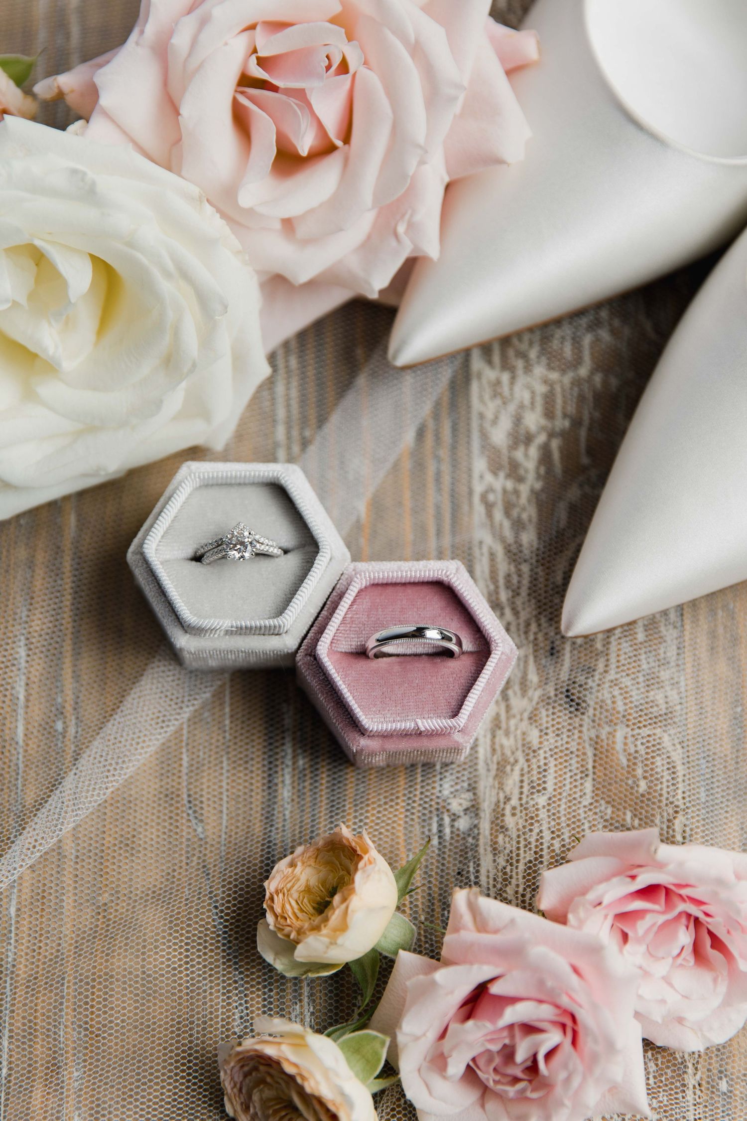 Wedding rings displayed in pink and gray hexagonal boxes with delicate roses on rustic wooden surface.