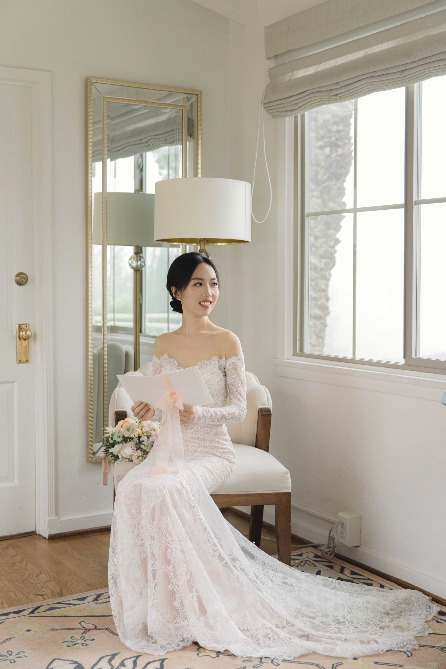 Elegant bride in lace off-shoulder wedding gown poses gracefully in a bright white room with floral bouquet.