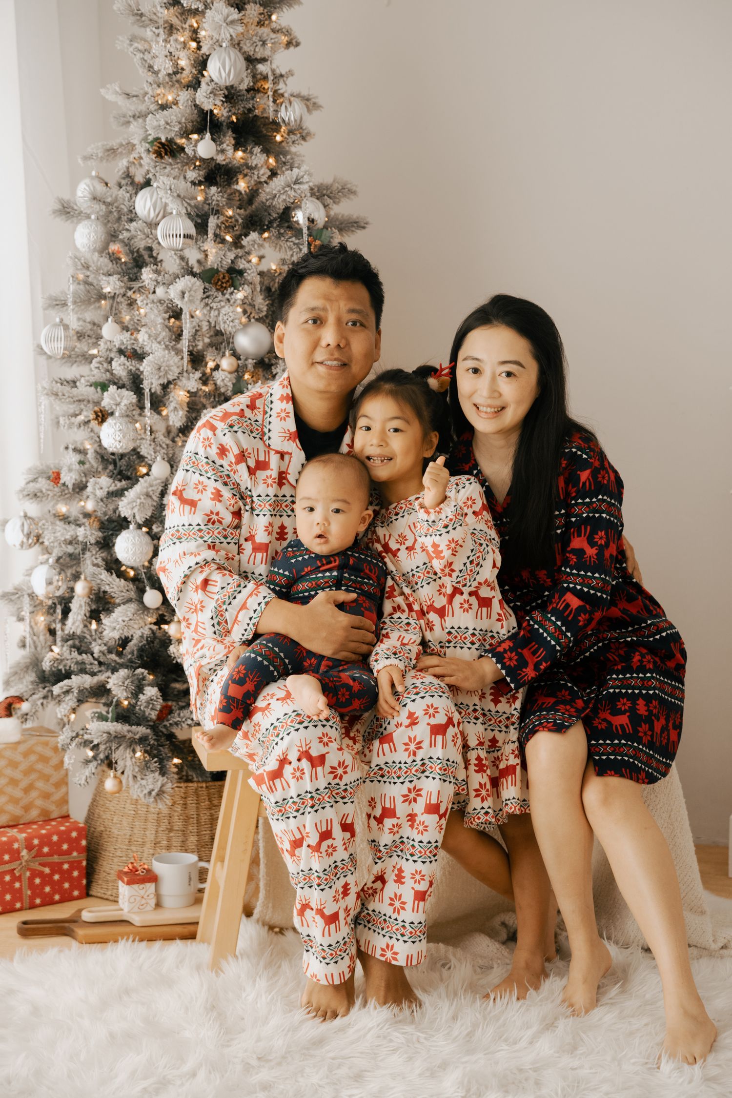 Family wearing matching holiday pajamas poses by decorated Christmas tree on white fluffy rug with wrapped presents.