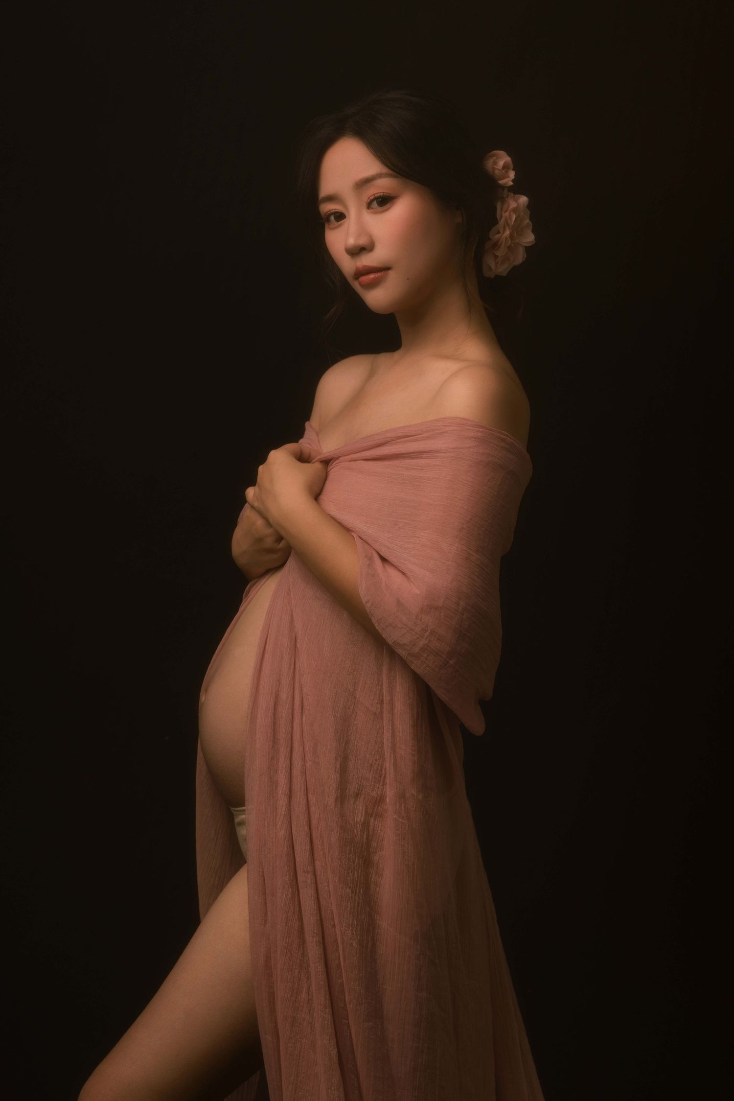 Moody maternity portrait with dusty rose fabric in dark studio setting.