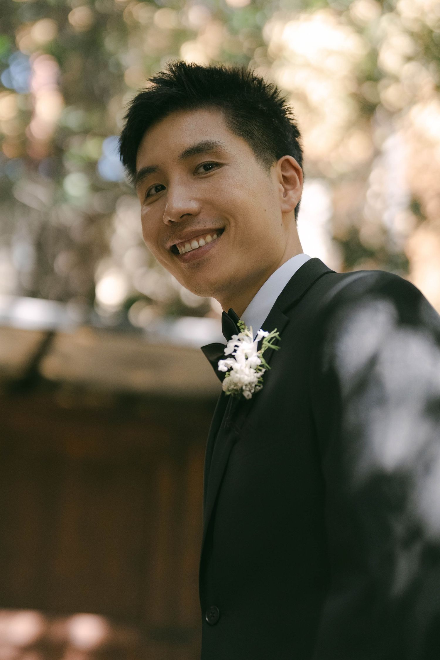 Groom wearing a black tuxedo with white boutonniere poses outdoors.
