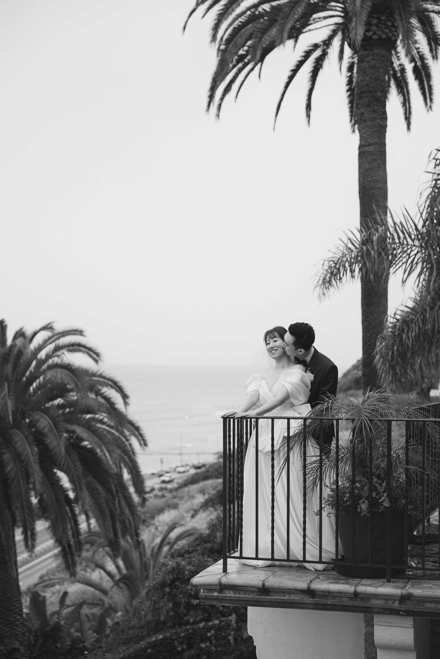 A couple shares an intimate moment on a balcony with palm trees and coastal views.