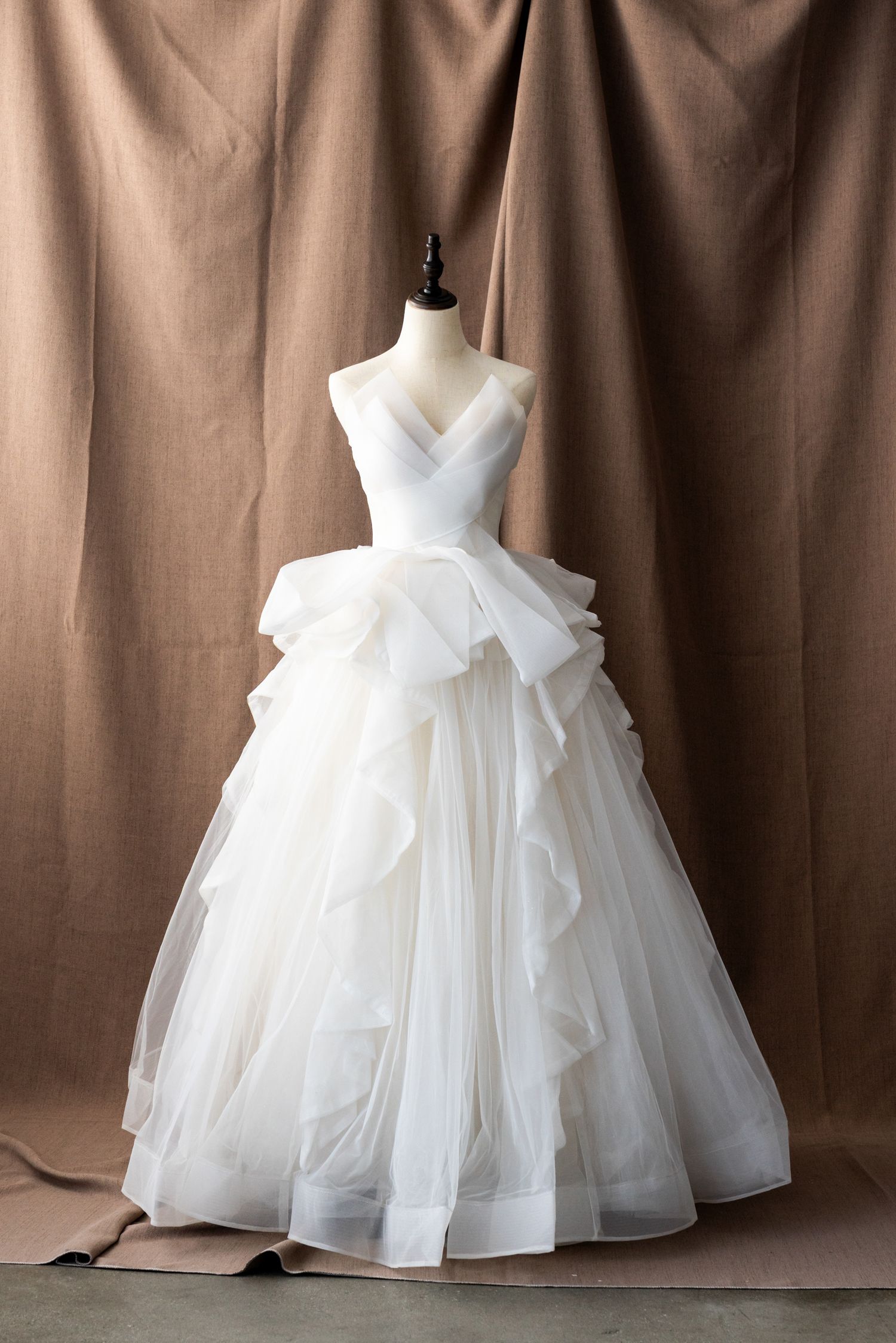 Full-skirted white wedding dress with ruffled tulle layers displayed against brown backdrop.