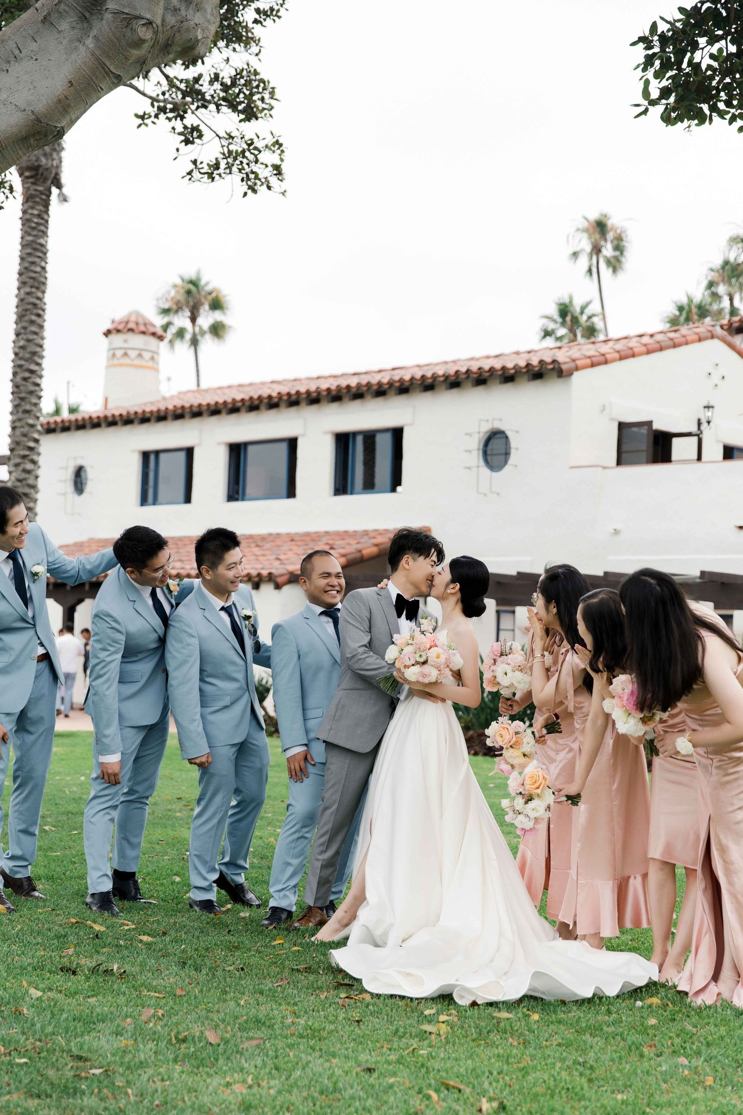 Wedding party in light blue suits and pink dresses poses on a lawn near a white Spanish-style building.