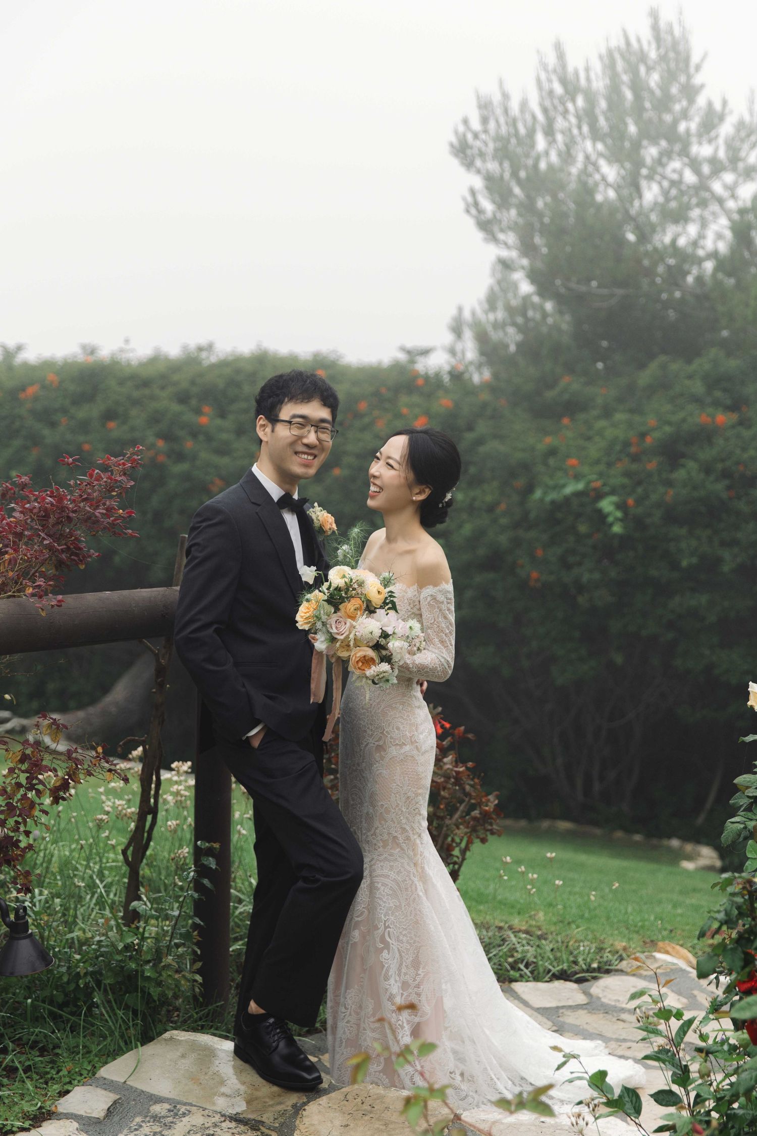 Joyful wedding couple laughs together in garden with blooming flowers and greenery.