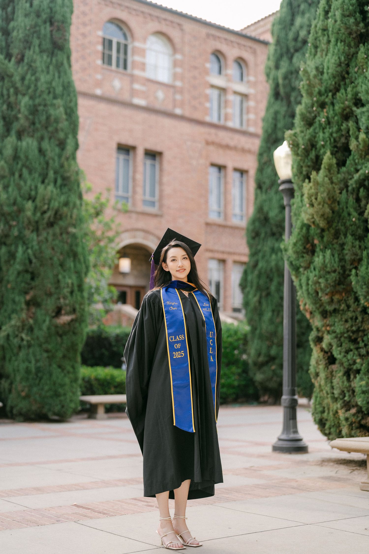 Graduate in academic regalia poses along cypress-lined pathway at USC campus.