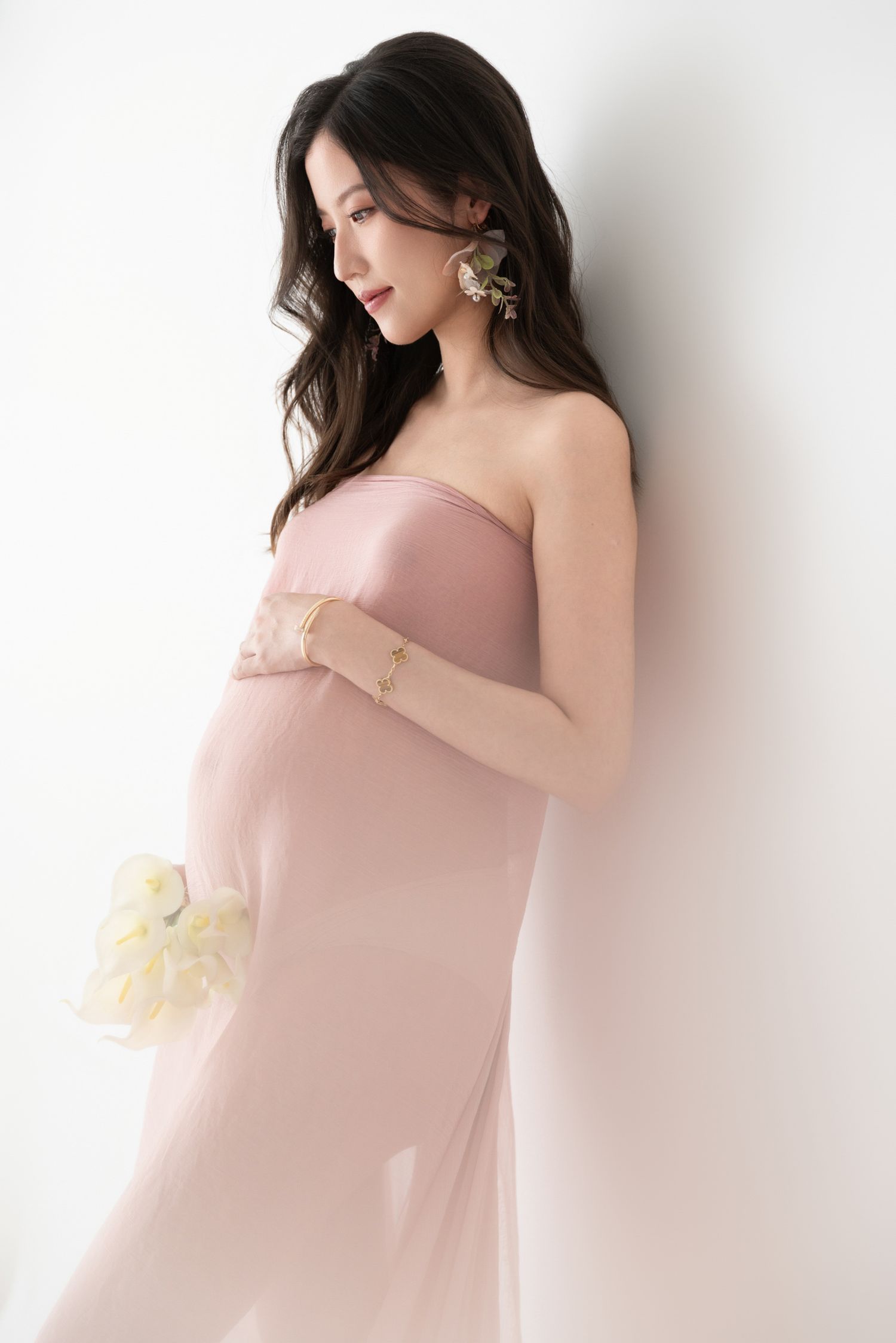 Elegant maternity portrait in soft pink dress against white backdrop.