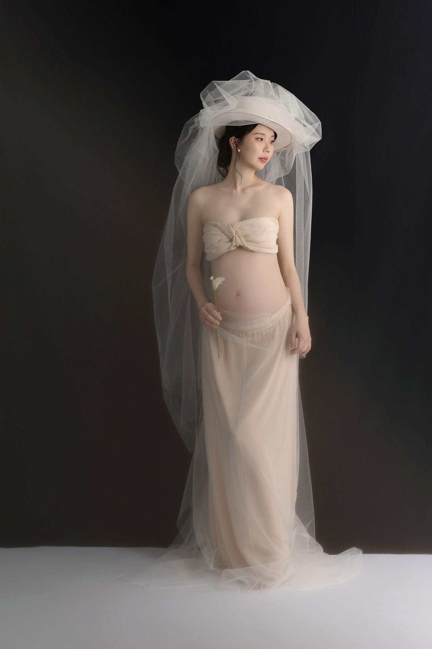 Dramatic maternity portrait with flowing tulle veil against dark background.