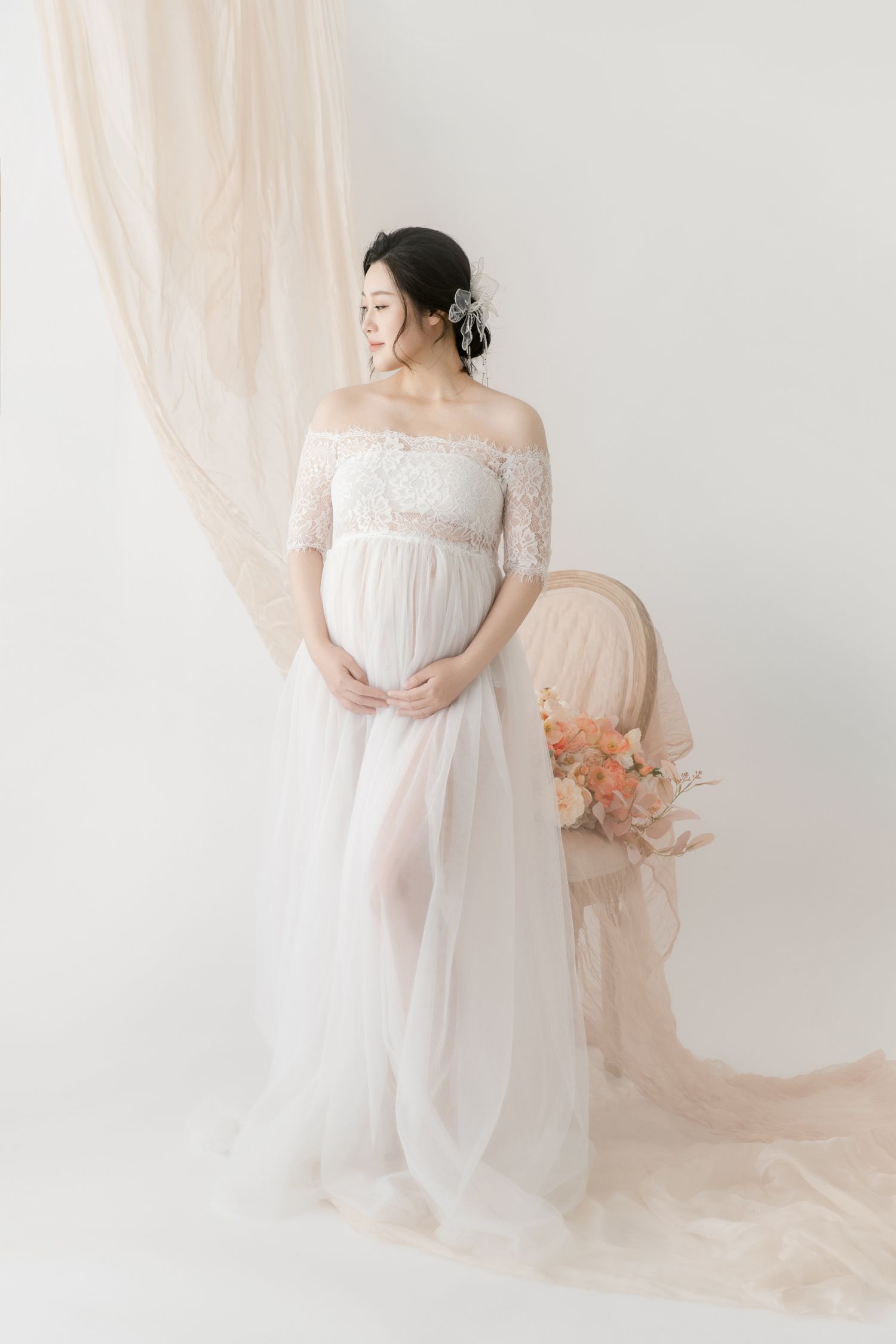 Elegant maternity session with flowing white gown and peach drapery in studio.