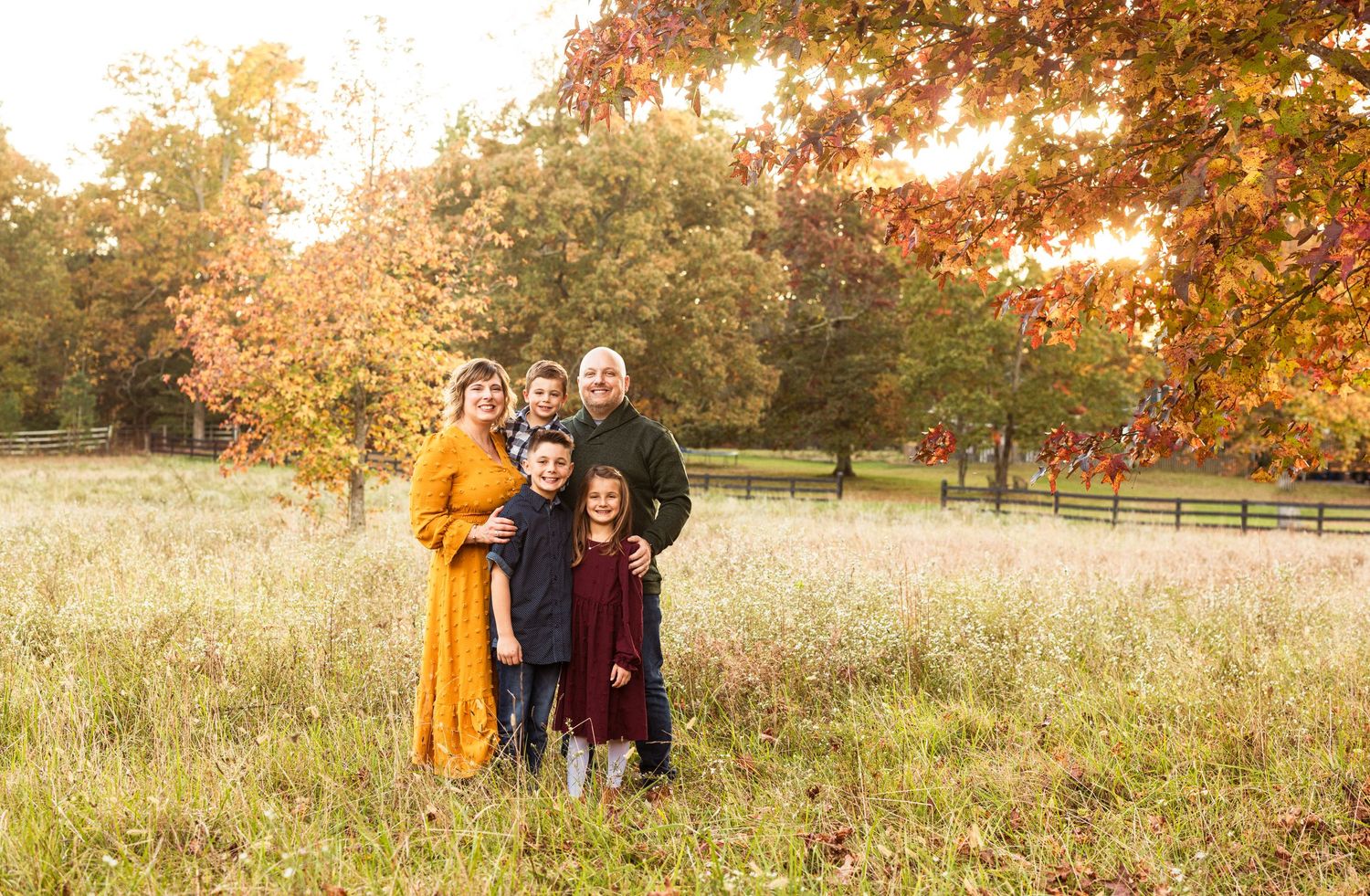 Family of five posing together in a sunlit autumn field surrounded by colorful fall foliage.