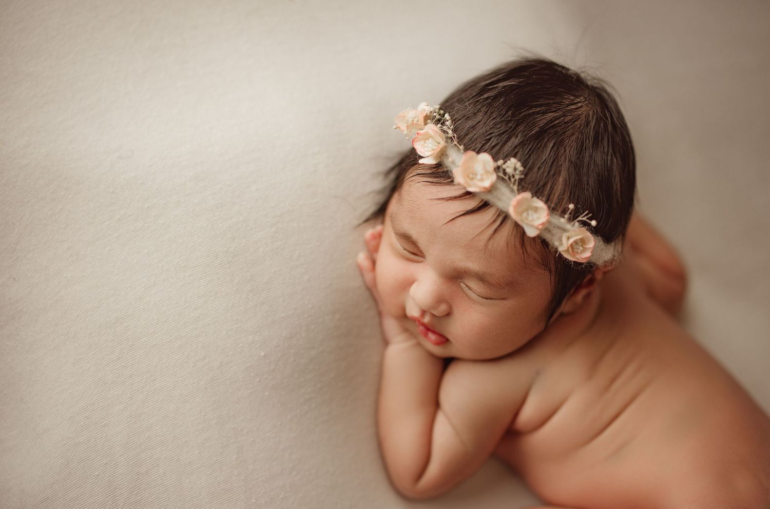 Sleeping newborn baby girl wearing a delicate floral crown, resting peacefully on a soft cream blanket.