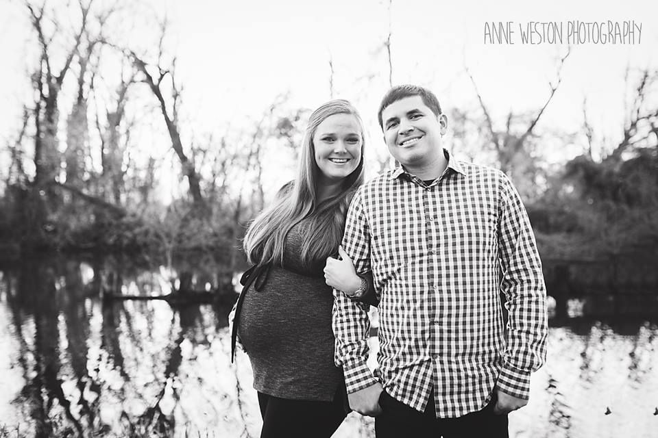 Black and white maternity photo of smiling couple standing together near a reflective pond outdoors.