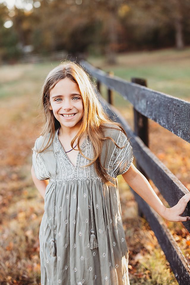 Young girl with long blonde hair smiling beside a wooden fence in a sage green dress outdoors. Richmond family photographer