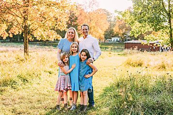 A family of five smiles together under golden autumn trees in a sunlit meadow.