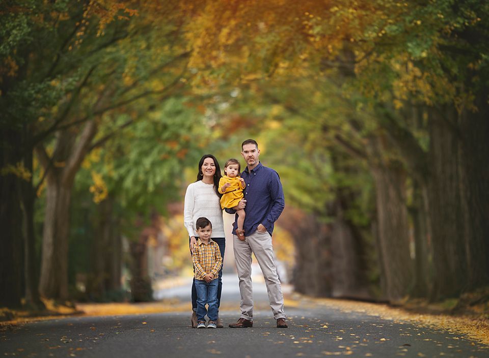 Family of four posing on a tree-lined road surrounded by vibrant autumn foliage in warm orange and yellow tones. Richmond family photography