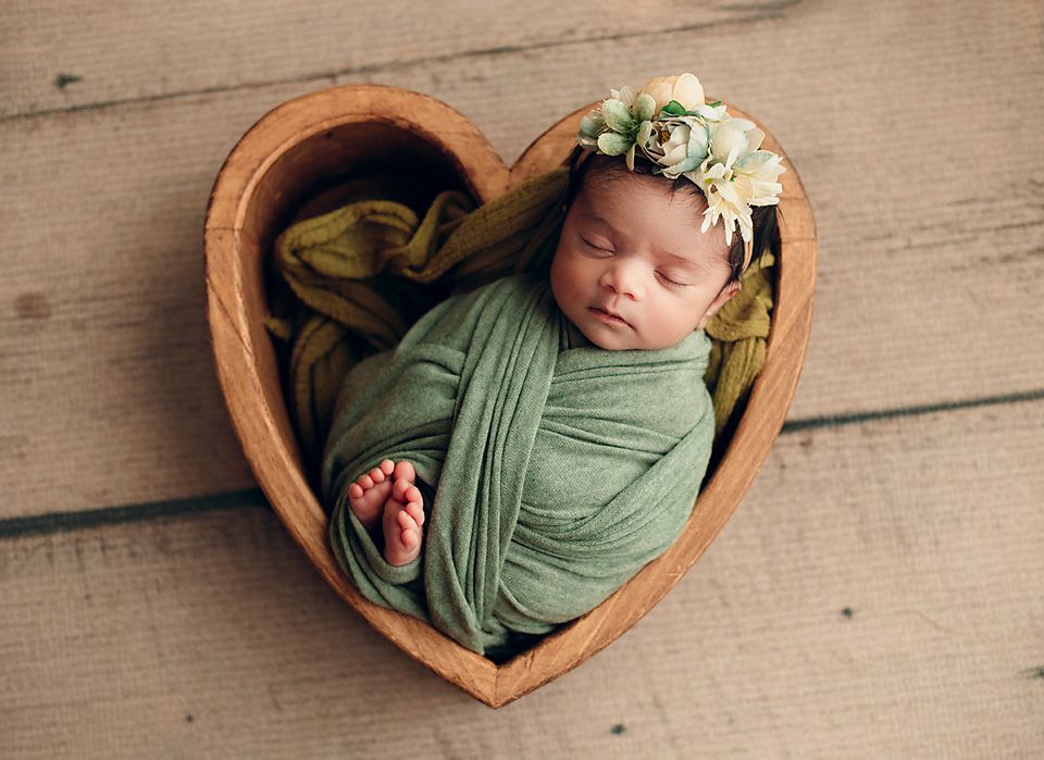 Sleeping newborn baby wrapped in green cloth with floral crown in heart-shaped wooden bowl.
