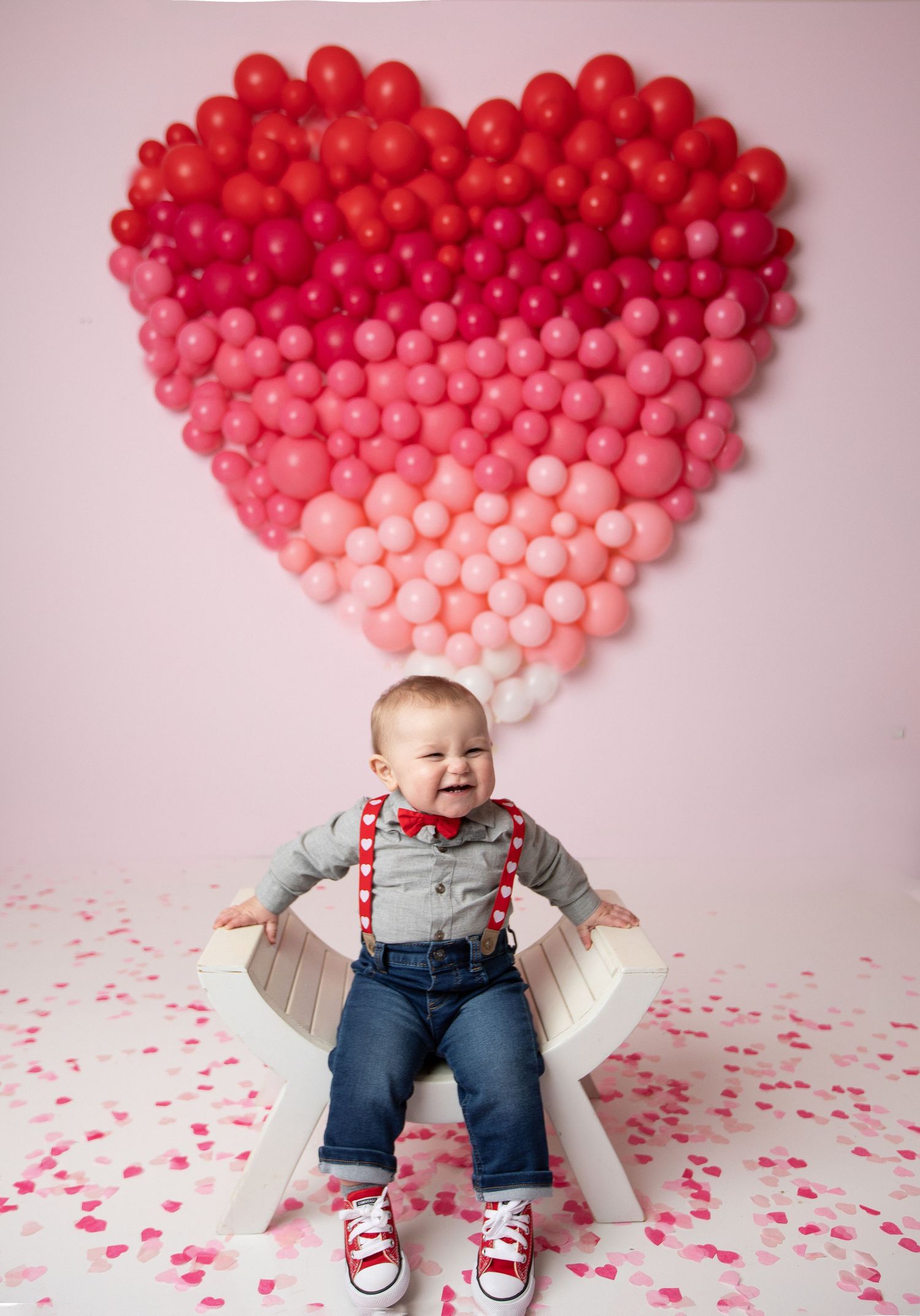 Smiling baby boy in suspenders and bow tie sits before a heart-shaped balloon wall for Valentine's Day.