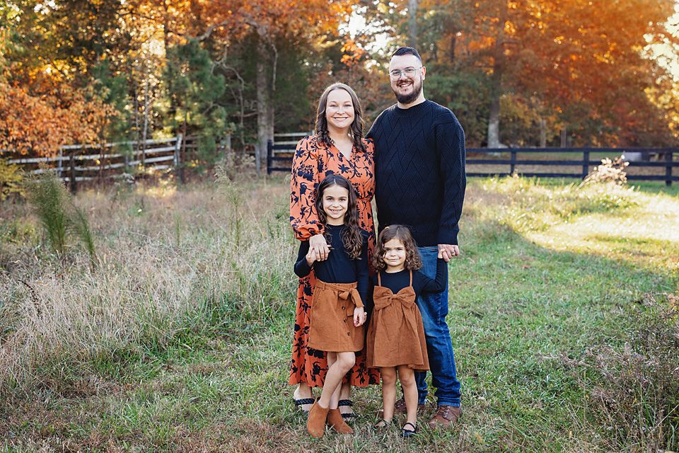 Family of four smiling outdoors in fall foliage with rustic fence backdrop.