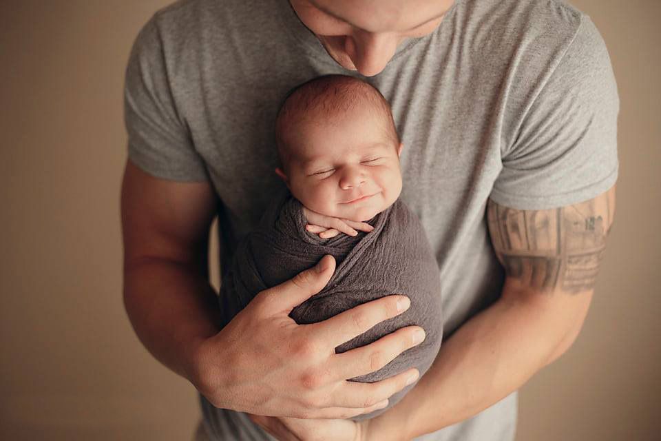 Father in gray t-shirt gently cradling sleeping swaddled newborn baby against chest.