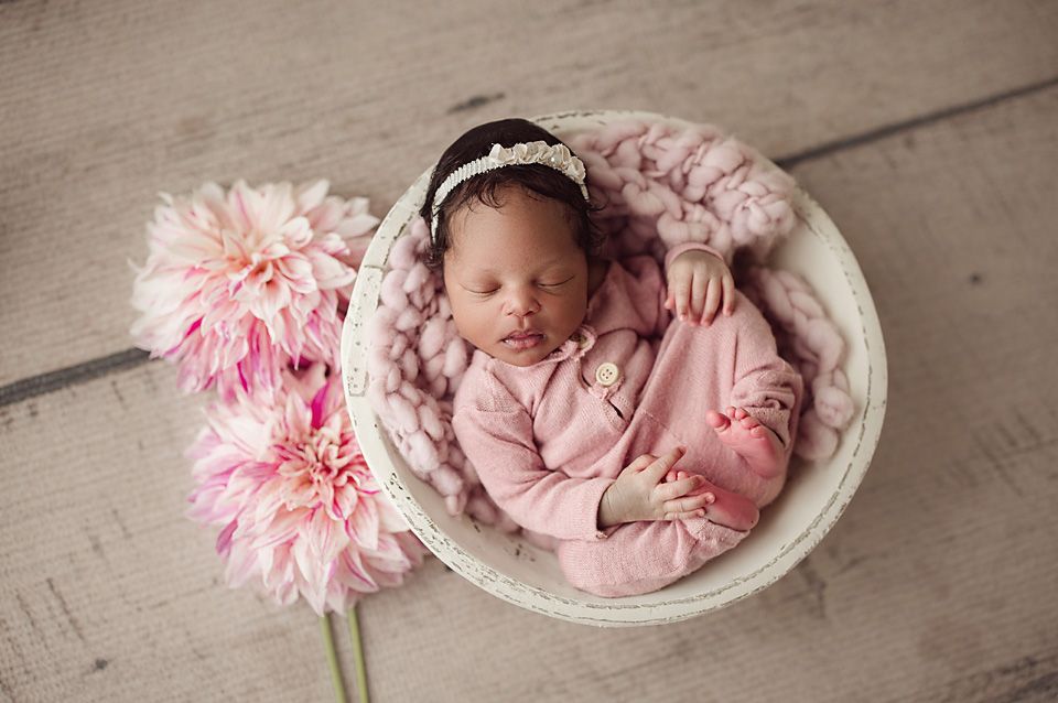 Newborn baby girl in pink outfit sleeping in a white basket surrounded by pink dahlia flowers.