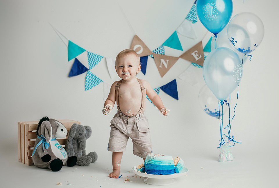 Baby boy celebrating first birthday standing by blue smash cake surrounded by blue balloons and toys. 1 year baby photography Petersburg
