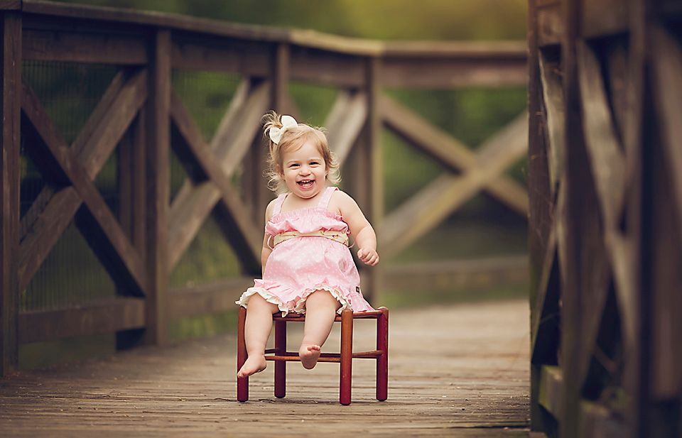 Smiling toddler girl in pink dress sitting on a small red chair on a wooden bridge.