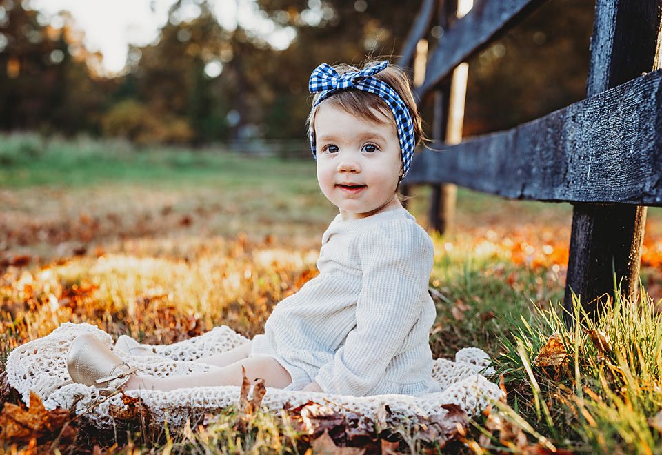 Cute baby girl in white dress and blue headband sits on a blanket among autumn leaves outdoors.