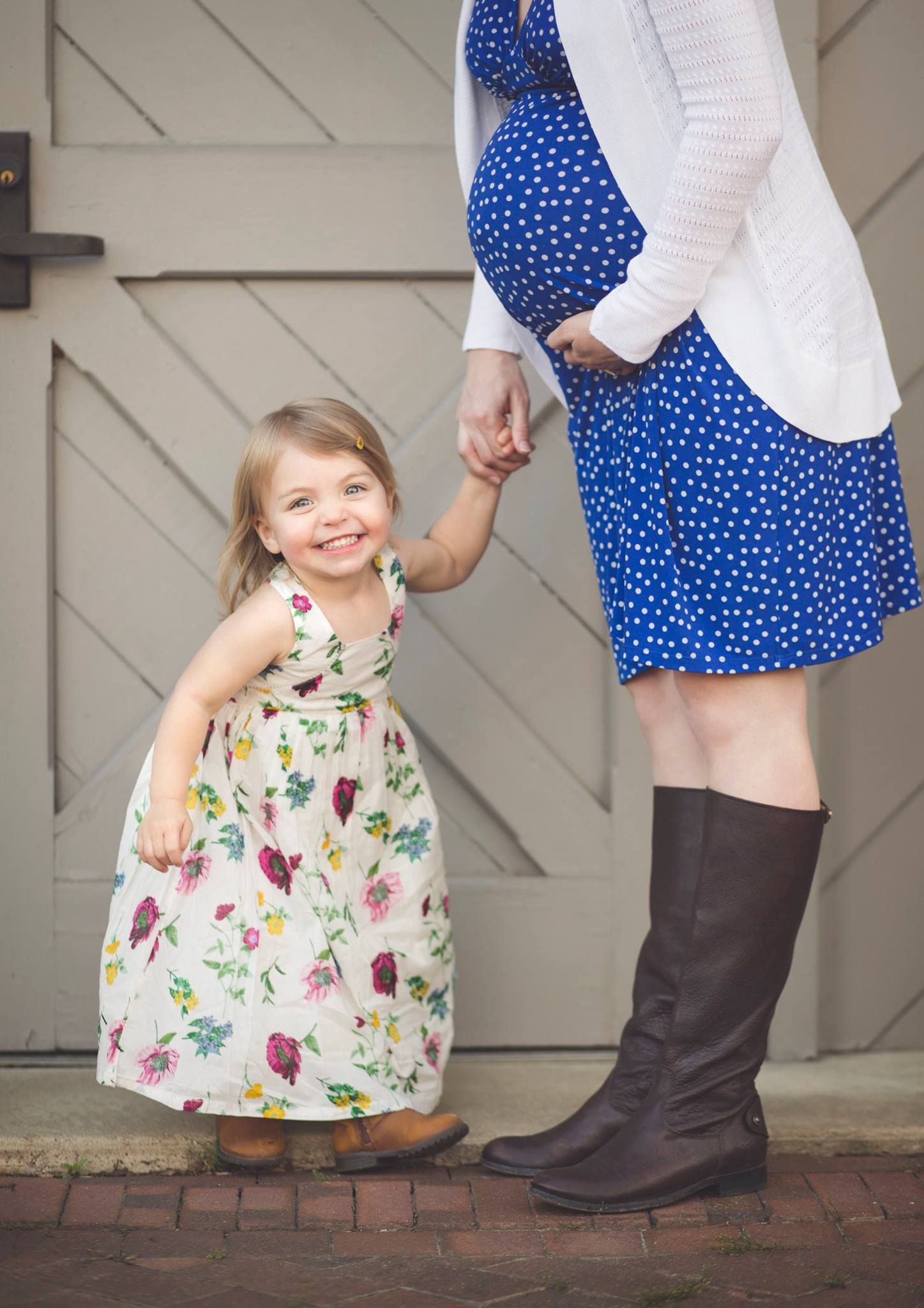 A smiling toddler girl in a floral dress holds hands with her pregnant mother in a blue polka dot dress and brown boots. Richmond maternity