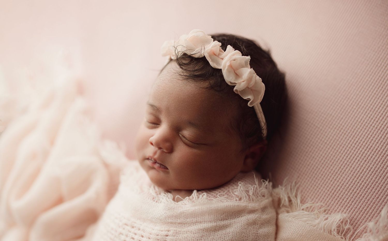 Sleeping newborn baby girl wrapped in white lace with a delicate pink floral headband on a soft pink background.