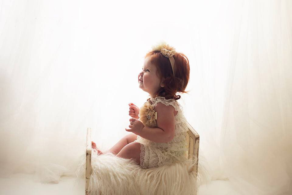 Adorable toddler girl in lace dress sitting on small bed holding stuffed animal in bright studio.