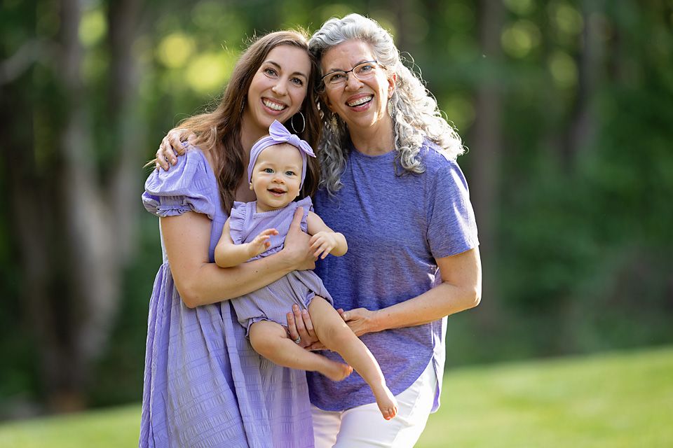 Two women in purple dresses smile outdoors holding a happy baby girl with a floral headband.