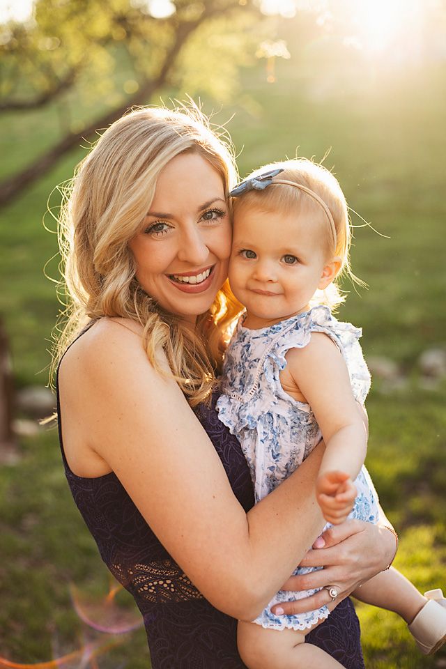 Glowing mother holding baby girl in blue floral dress during golden hour outdoor portrait session.