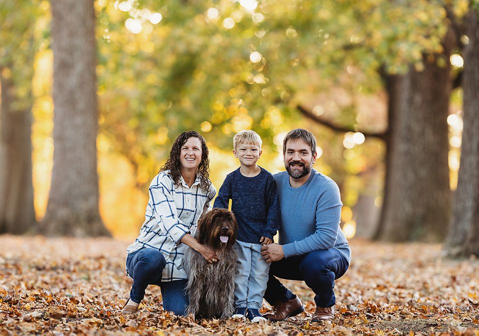 Family of three with dog posing among autumn leaves and trees.