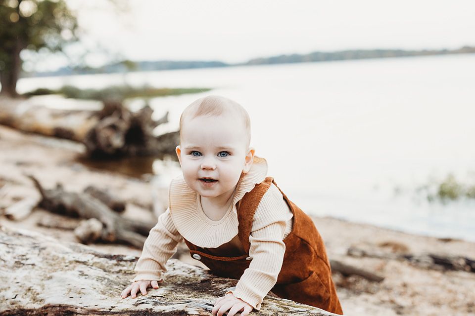 Adorable baby in rust overalls crawls on rocks near a serene lakeside during an outdoor photo session Richmond family photography Hopewell