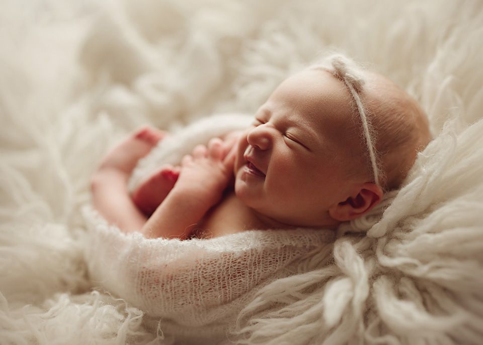 Adorable newborn baby girl resting on soft white fur blanket, wearing a tiny white headband, with a gentle smile.