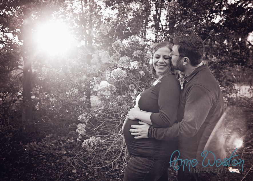 Black and white maternity photo of a smiling pregnant woman being embraced by her partner outdoors among blooming trees.. Richmond VA