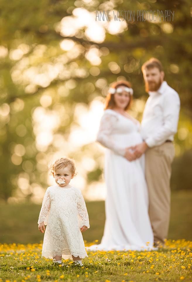 Pregnant woman in white lace gown with bearded husband outdoors, toddler girl in white dress with pacifier in foreground. Maternity photo