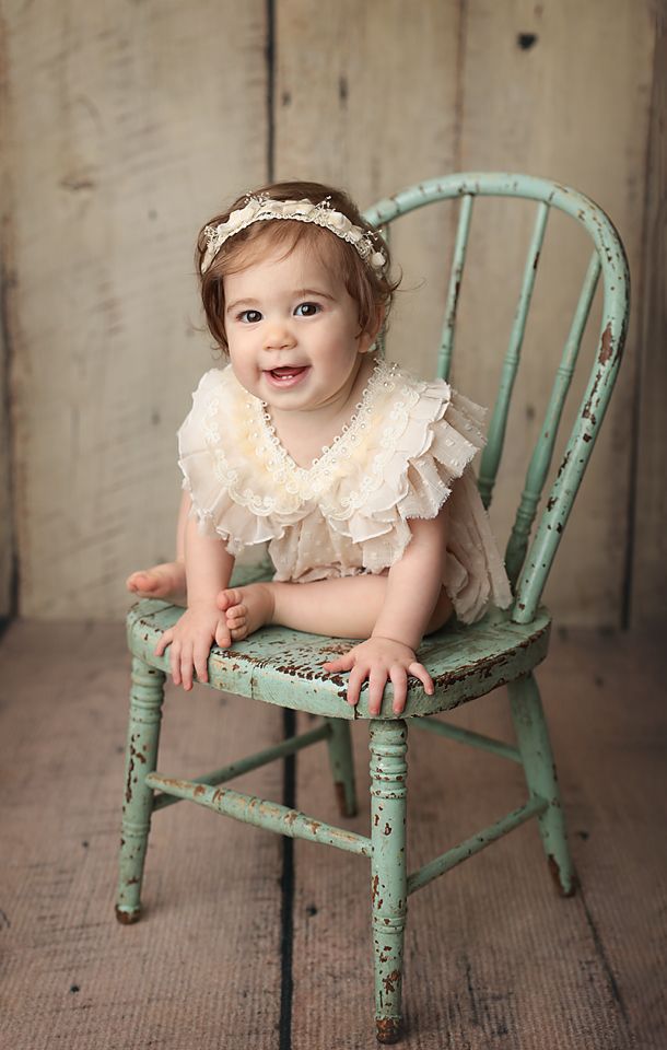 Smiling baby girl in lace romper and floral headband sitting on vintage teal distressed wooden chair.