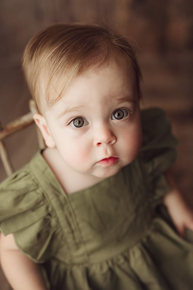 Close-up portrait of a baby girl in a green dress with big gray eyes on a dark background.