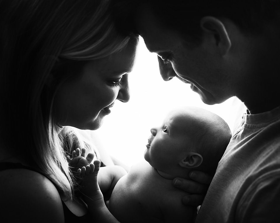 Black and white photo of two parents lovingly gazing at their newborn baby held between them.