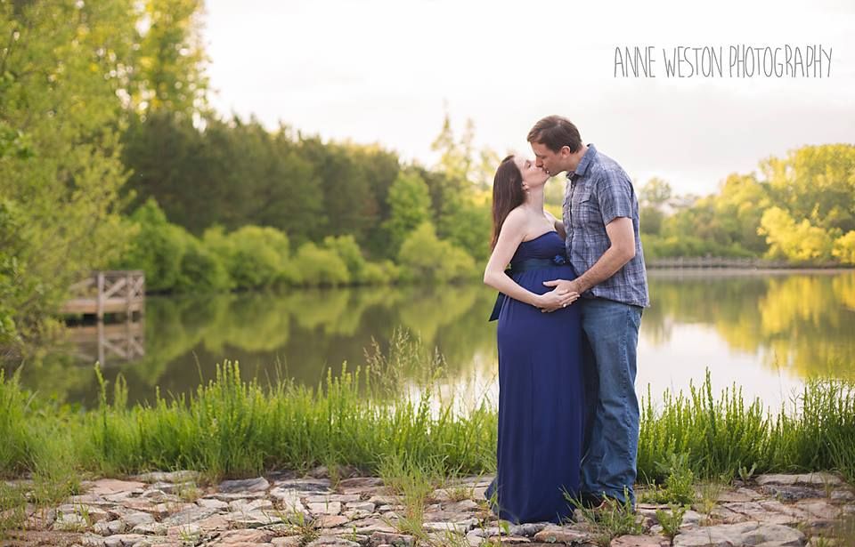 Couple sharing a tender kiss by a serene lake, woman in blue maternity gown, lush green trees in background. Maternity Photography session