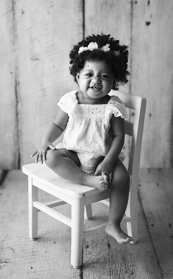 Adorable toddler girl with curly hair and flower crown sitting on white chair in black and white.