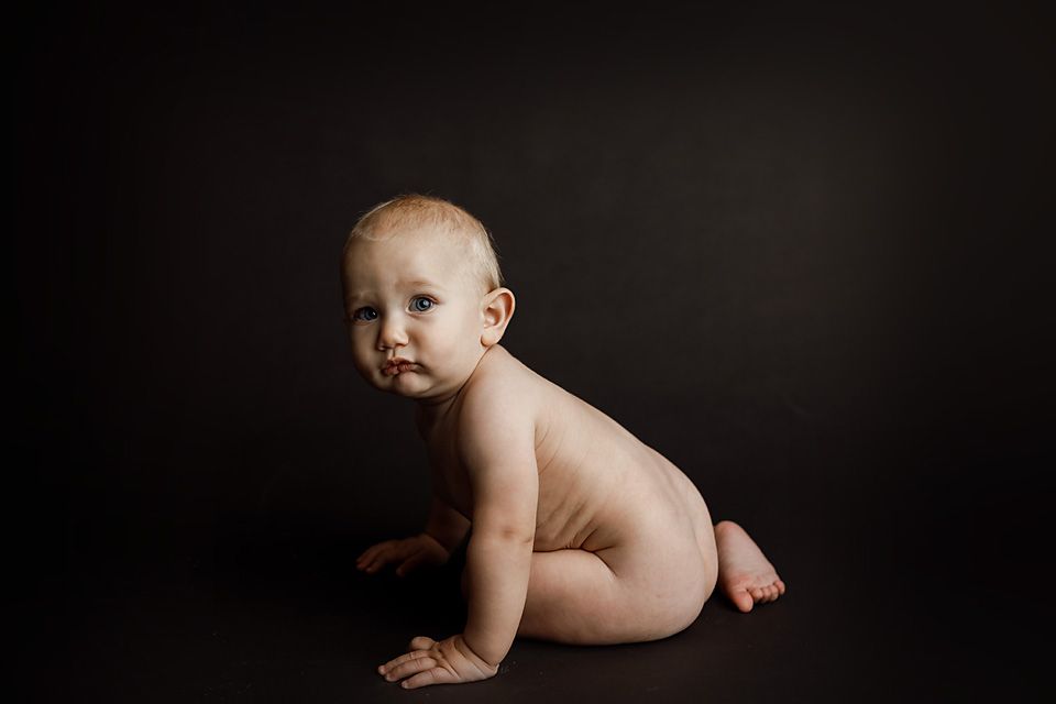 Naked baby crawls on a dark background, looking over shoulder in a minimalist studio portrait.