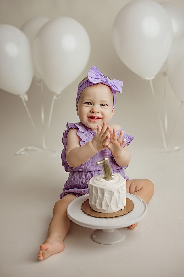 Joyful baby girl in purple romper claps hands over smash cake surrounded by white balloons.