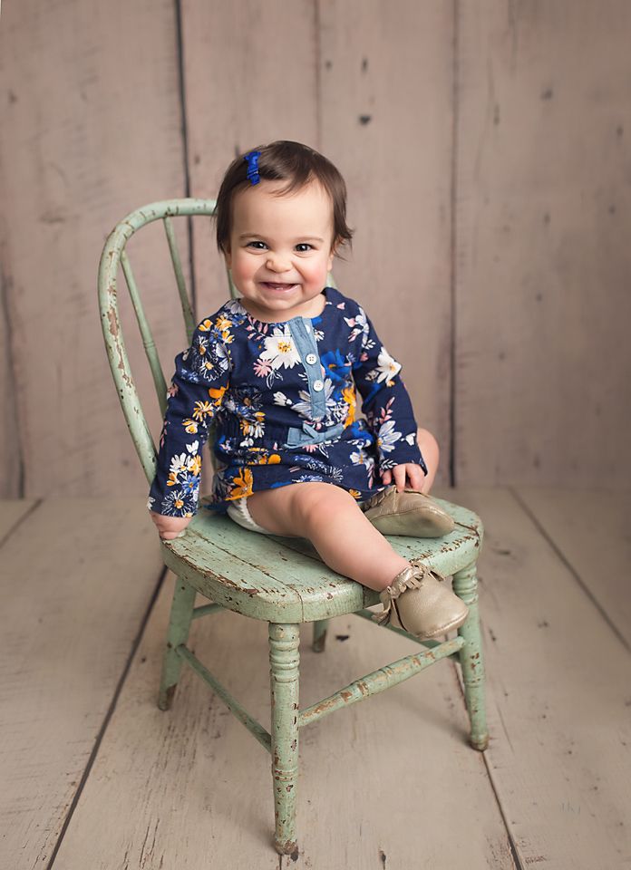 Smiling baby girl in floral navy dress sitting on a vintage mint green wooden chair.