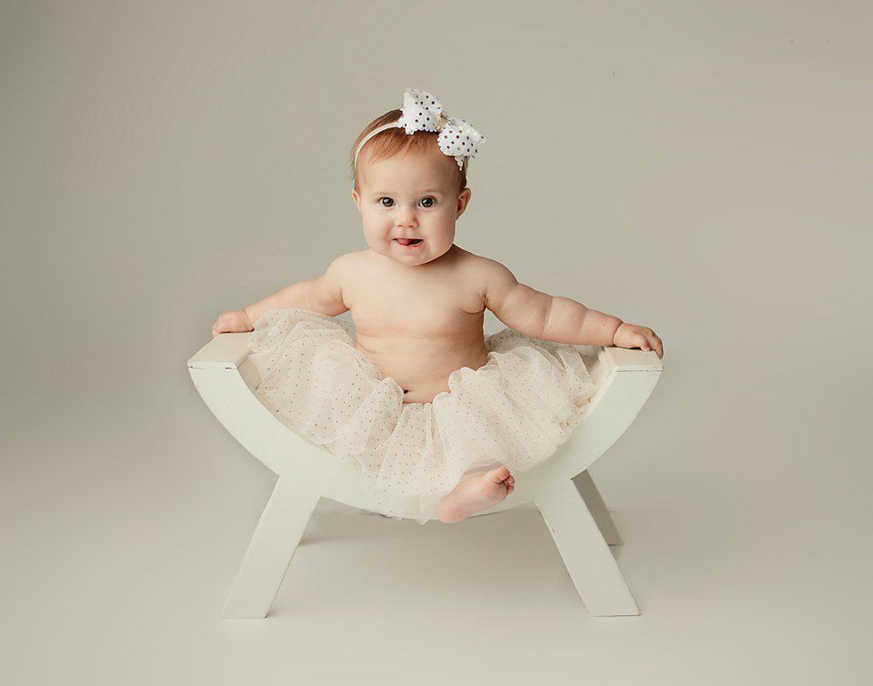 Smiling baby girl in cream tutu skirt and bow headband sitting in white wooden prop chair. baby photography Chesterfield